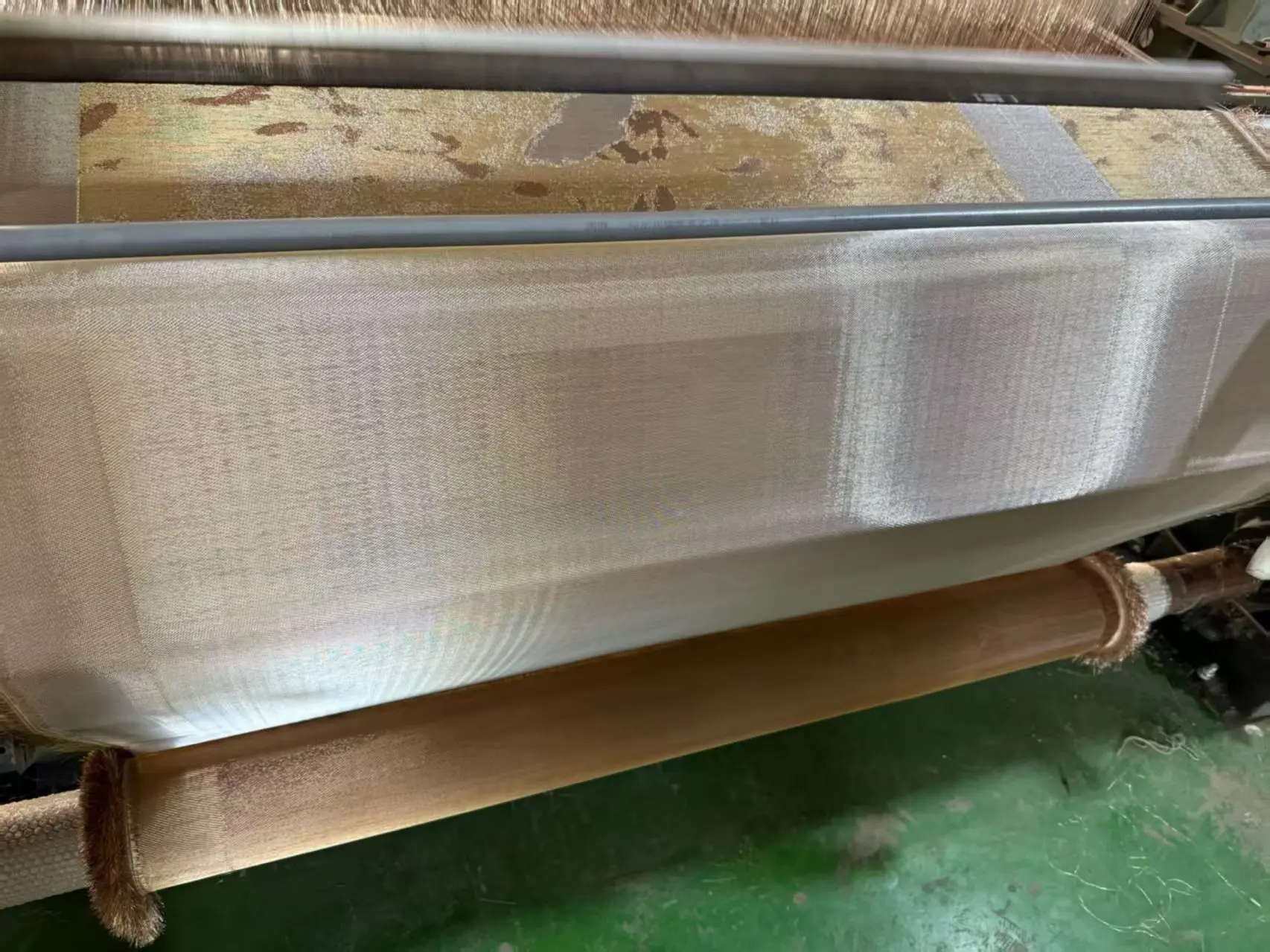 Close-Up Of A Textile Loom Producing A Light, Shimmering Jacquard Fabric, Showing Warp Threads, Rollers, And Woven Cloth In Progress.